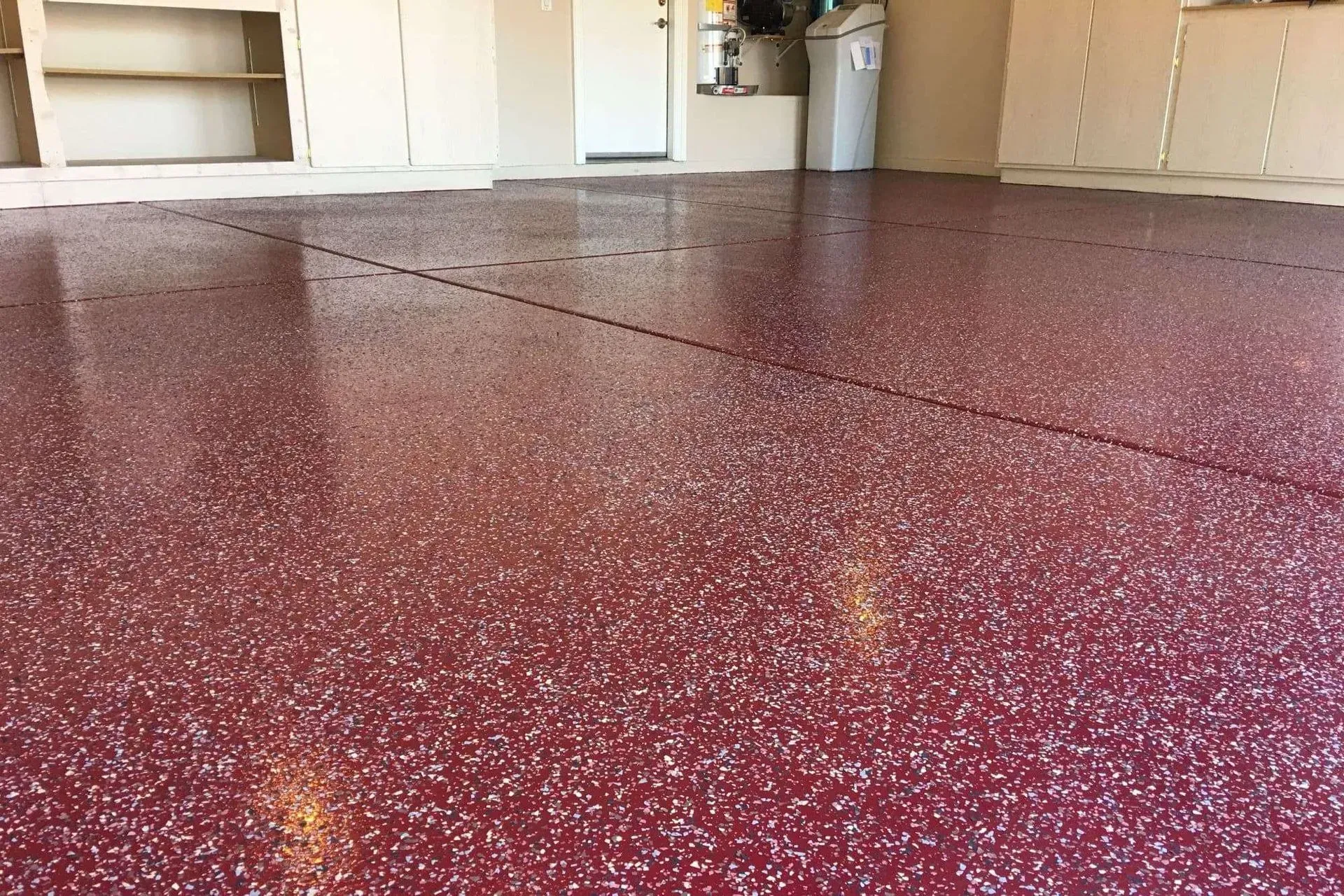 Red epoxy-coated garage floor with white flecks, reflecting light. Shelves and a white door are visible in the background.