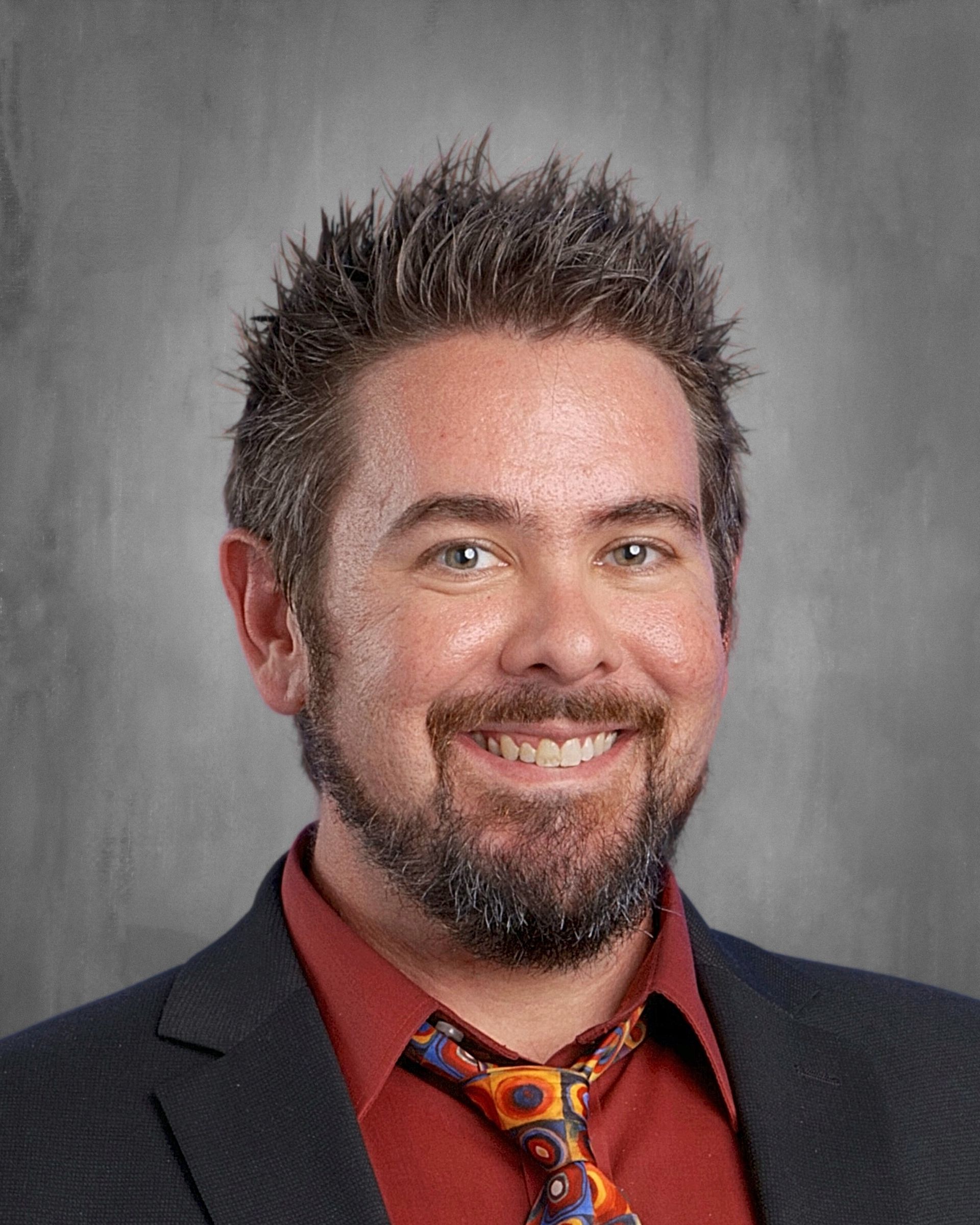 Man with spiky hair, wearing a red shirt, tie, and blazer, smiling at the camera.