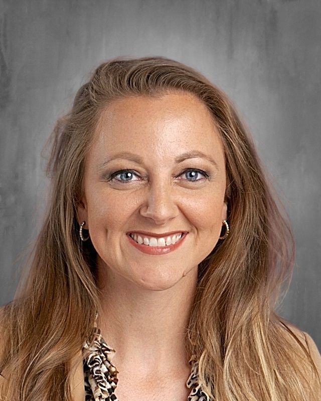 Woman with long blonde hair, smiling, wearing hoop earrings, against a gray background.