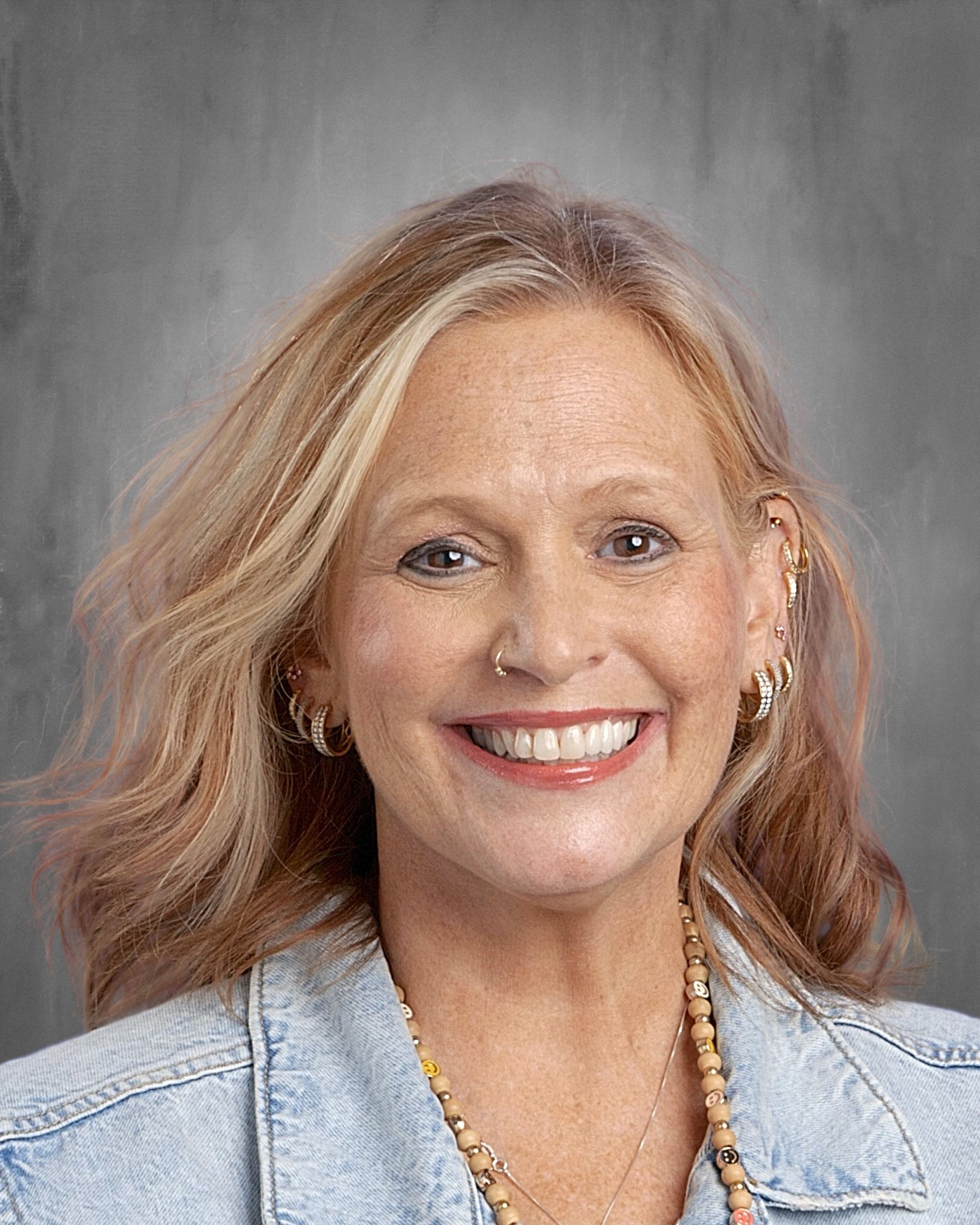 Smiling woman with blonde hair wearing a denim jacket and leopard print top; gold hoop earrings and necklace.