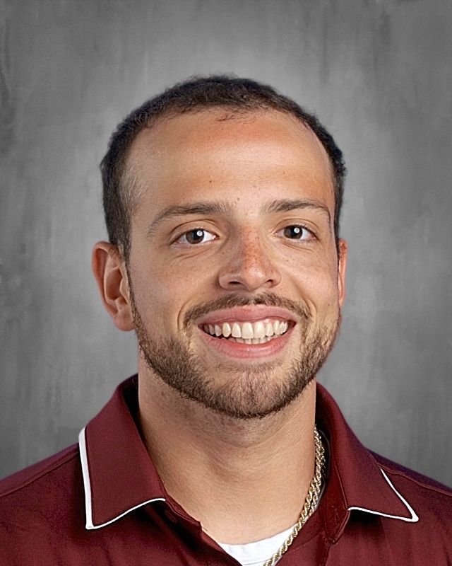 A young man with brown curly hair and a beard, smiling. He wears a pink polo shirt and has a gold chain.