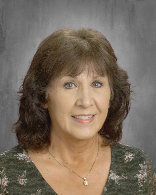Woman with shoulder-length brown hair smiles at the camera. She wears a floral green shirt and a pendant.