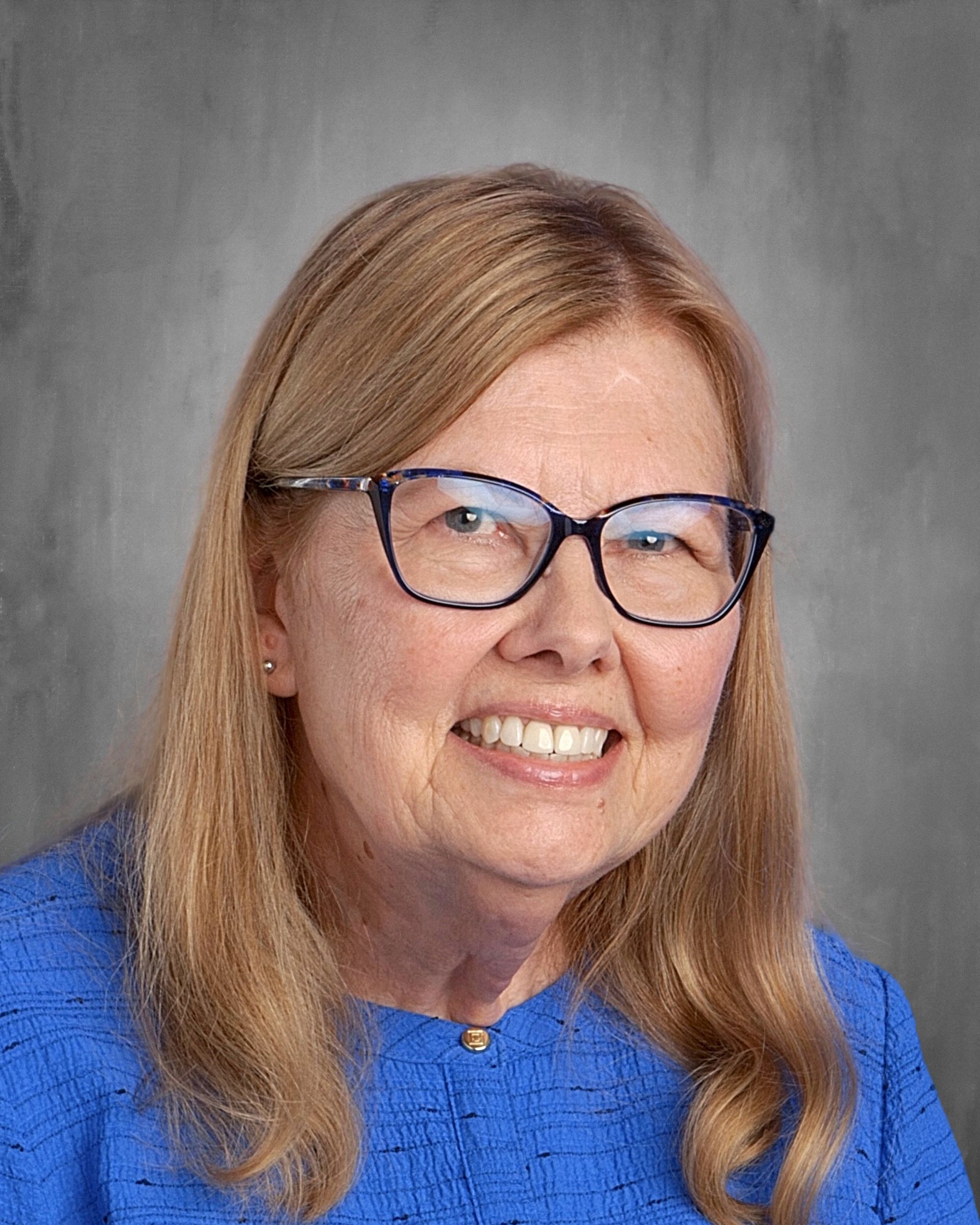 Woman with blonde hair and glasses smiles; wearing a patterned shirt against a grey backdrop.