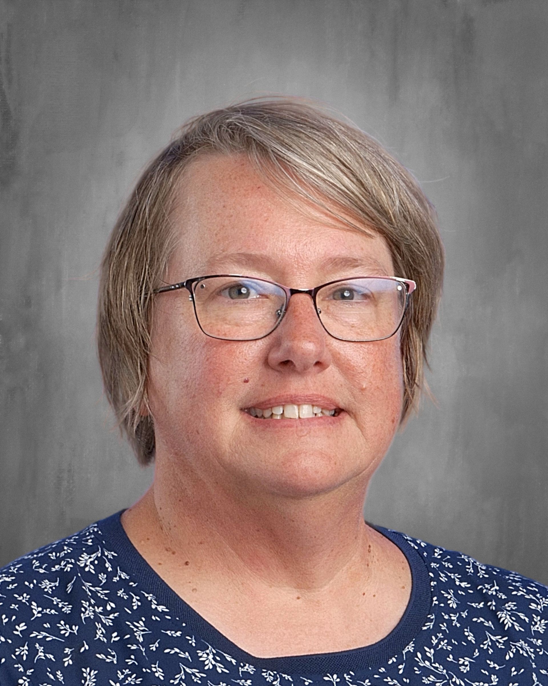 Woman with short blonde hair, glasses, smiling. Wearing a blue floral top, against a gray backdrop.