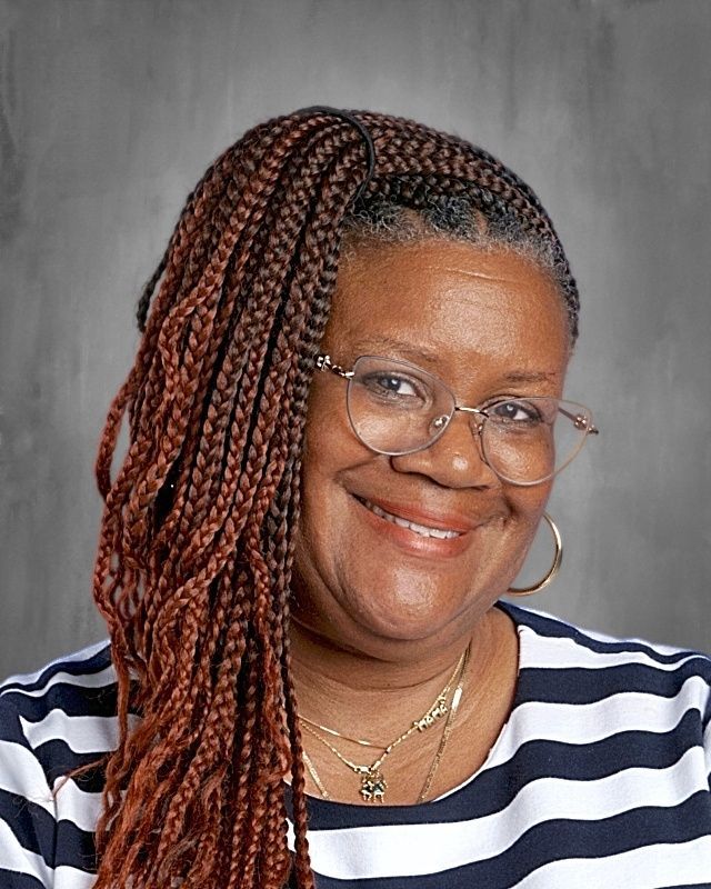 Woman with glasses and natural hair smiling, wearing a blue shirt and beaded necklace, set against a gray background.