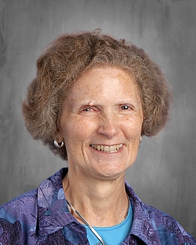 Woman with curly brown hair, smiling, wearing purple sweater and necklace, against a gray background.