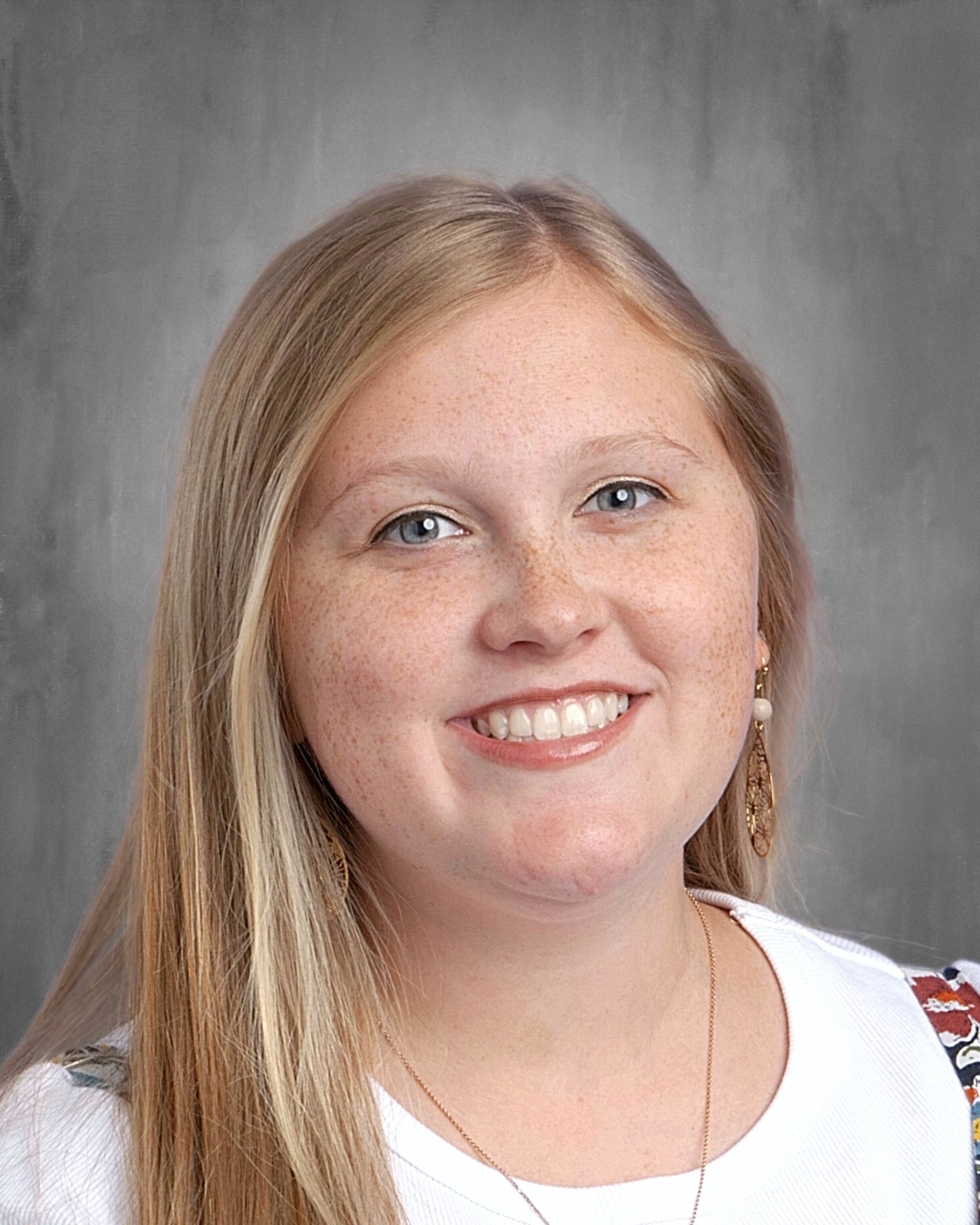 Woman with blonde hair smiles, wearing a purple shirt, against a gray background.