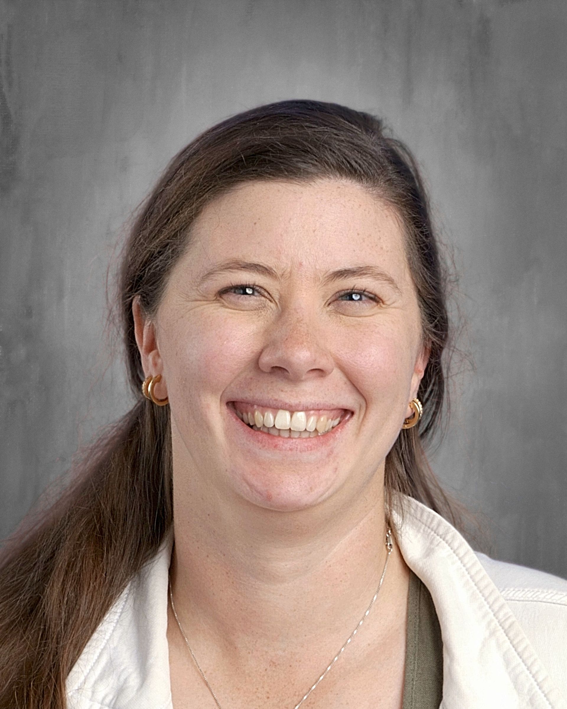 Woman with long brown hair, smiling, wearing white jacket and olive green shirt, gray background.