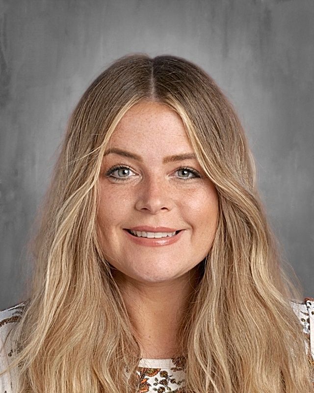 Woman with long, wavy brown hair smiles. She wears a colorful top against a gray backdrop.