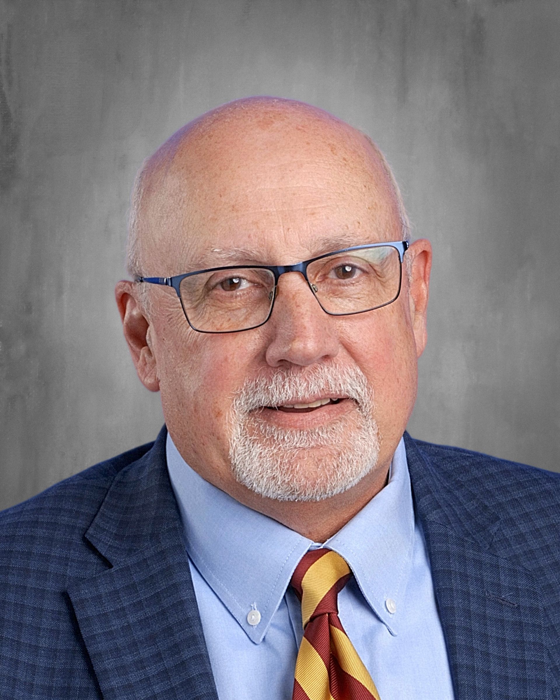 Man with glasses, smiling, wearing a suit and tie. Gray background.