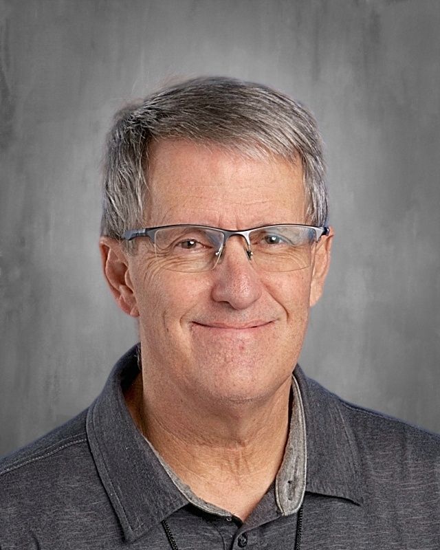 Older man with glasses and a blue shirt smiles at the camera, against a gray background.