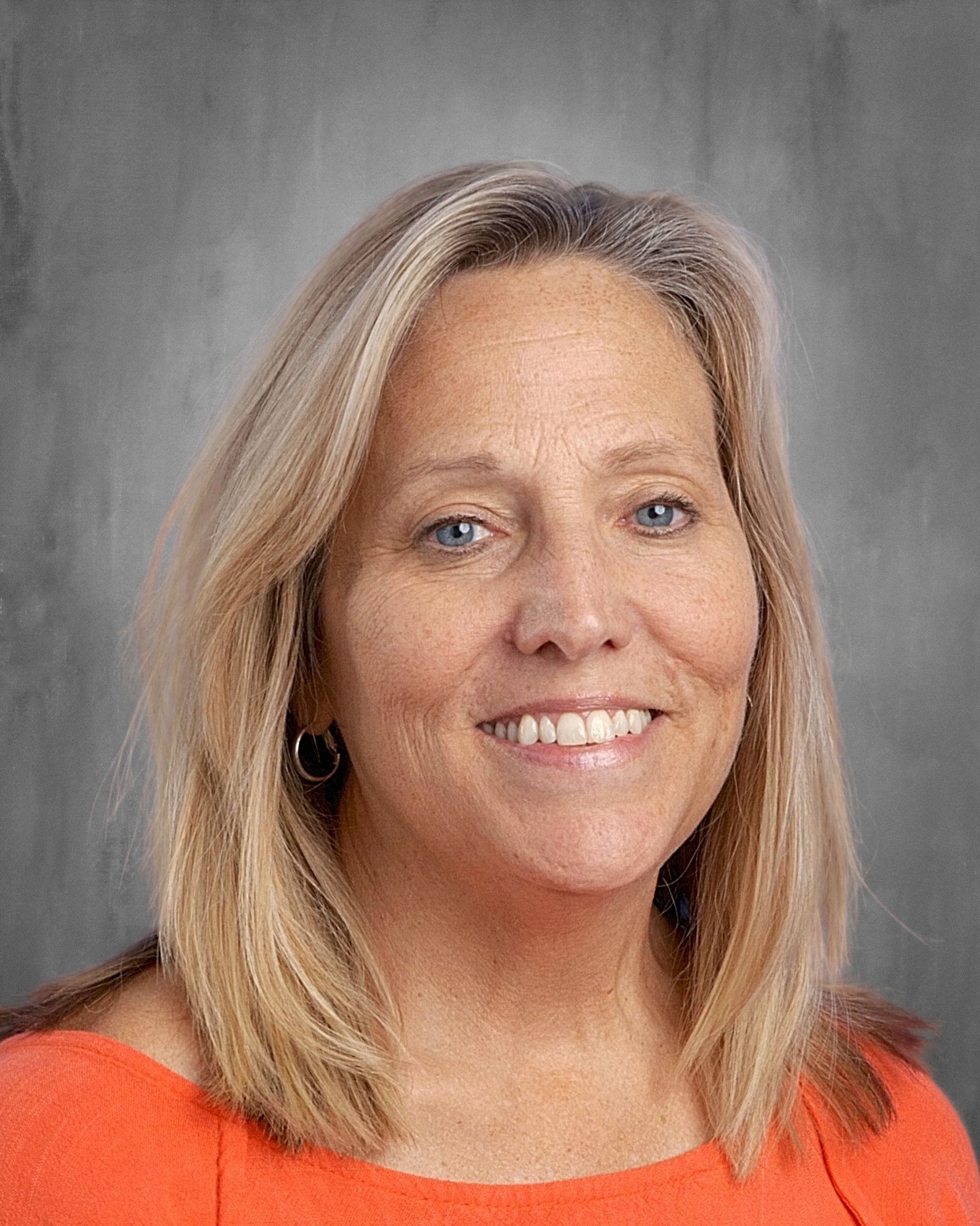 Woman with blonde hair, smiling, wearing a light blue shirt. Gray backdrop.