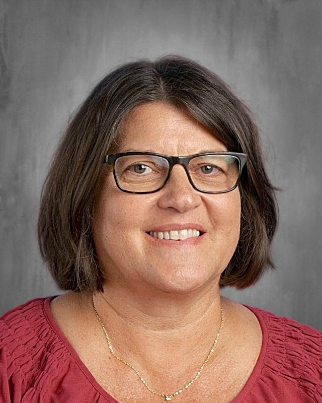 Woman with glasses smiling, wearing a blue floral shirt, gray backdrop.