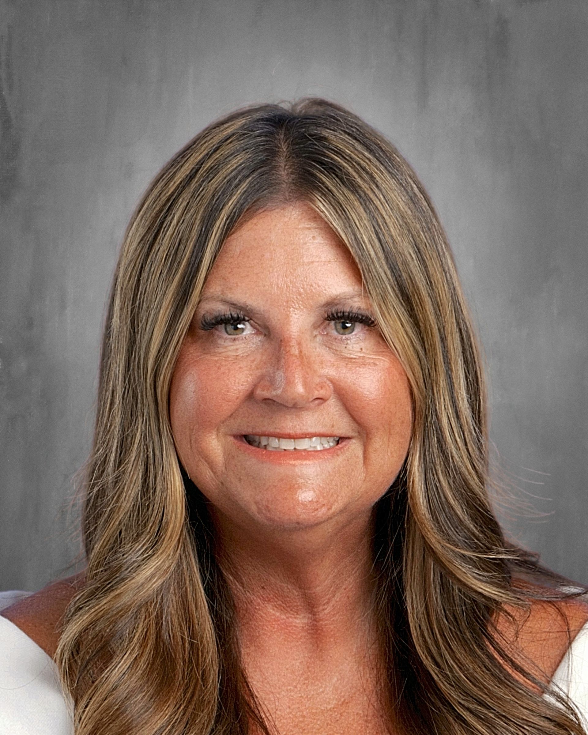Woman with long brown hair, smiling, wearing a white top, against a gray background.