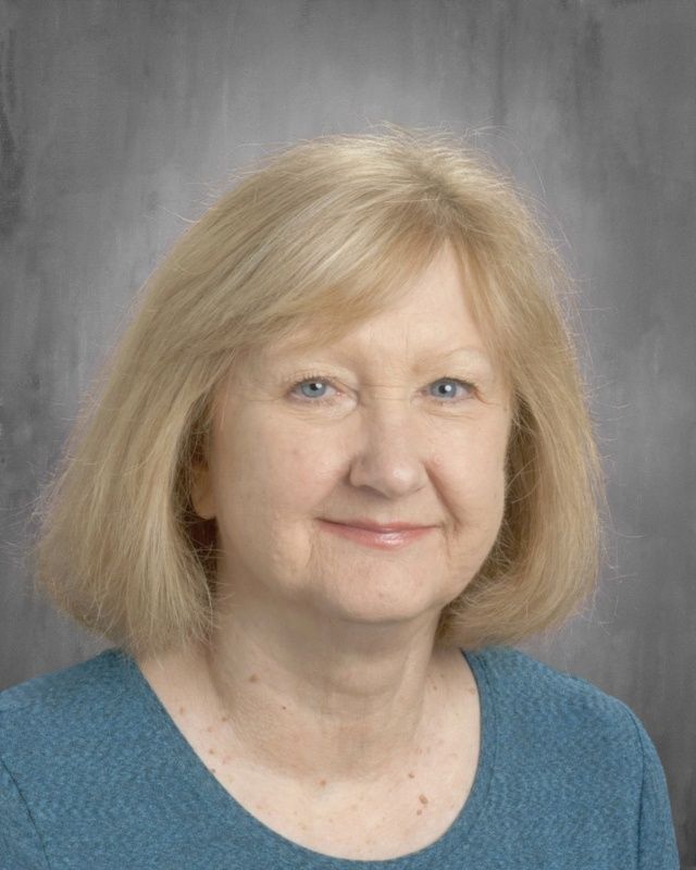 Blonde woman with blue eyes and a slight smile, wearing a blue sweater, against a gray backdrop.