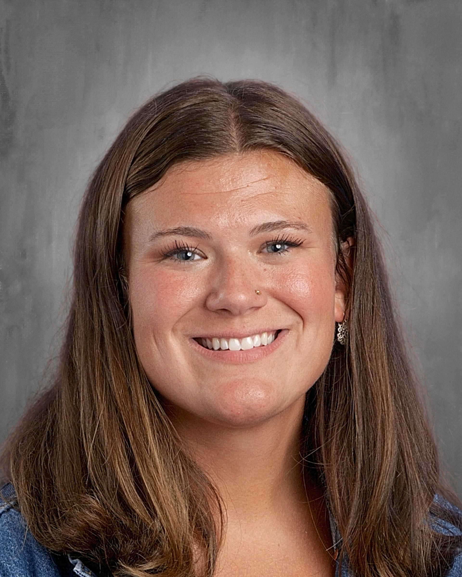 Woman with blonde hair smiles, wearing a green floral top, gold necklace, and drop earrings. Gray backdrop.