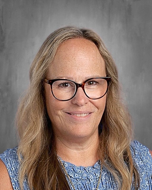 Woman with blonde hair and glasses, smiling, looking at the camera. Against a gray background.