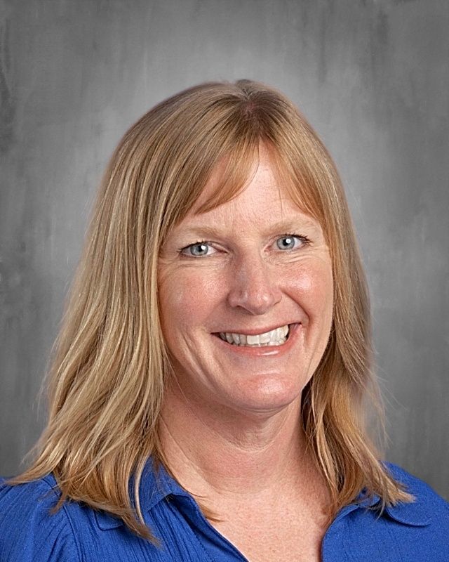 Blond woman with fair skin and a friendly smile, wearing a green top, against a gray background.