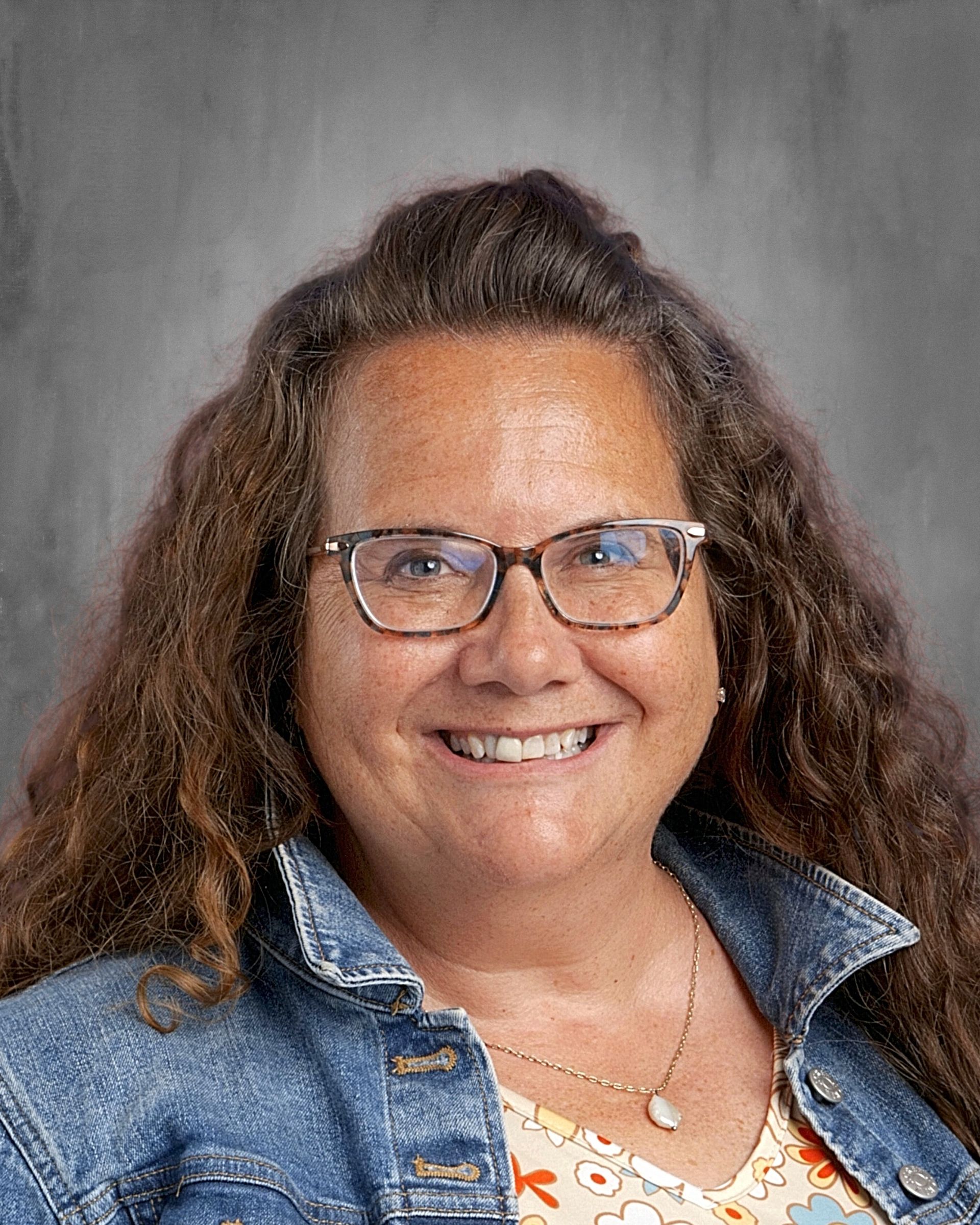 Woman with glasses smiles, wearing a green shirt and beaded necklace, against a grey background.