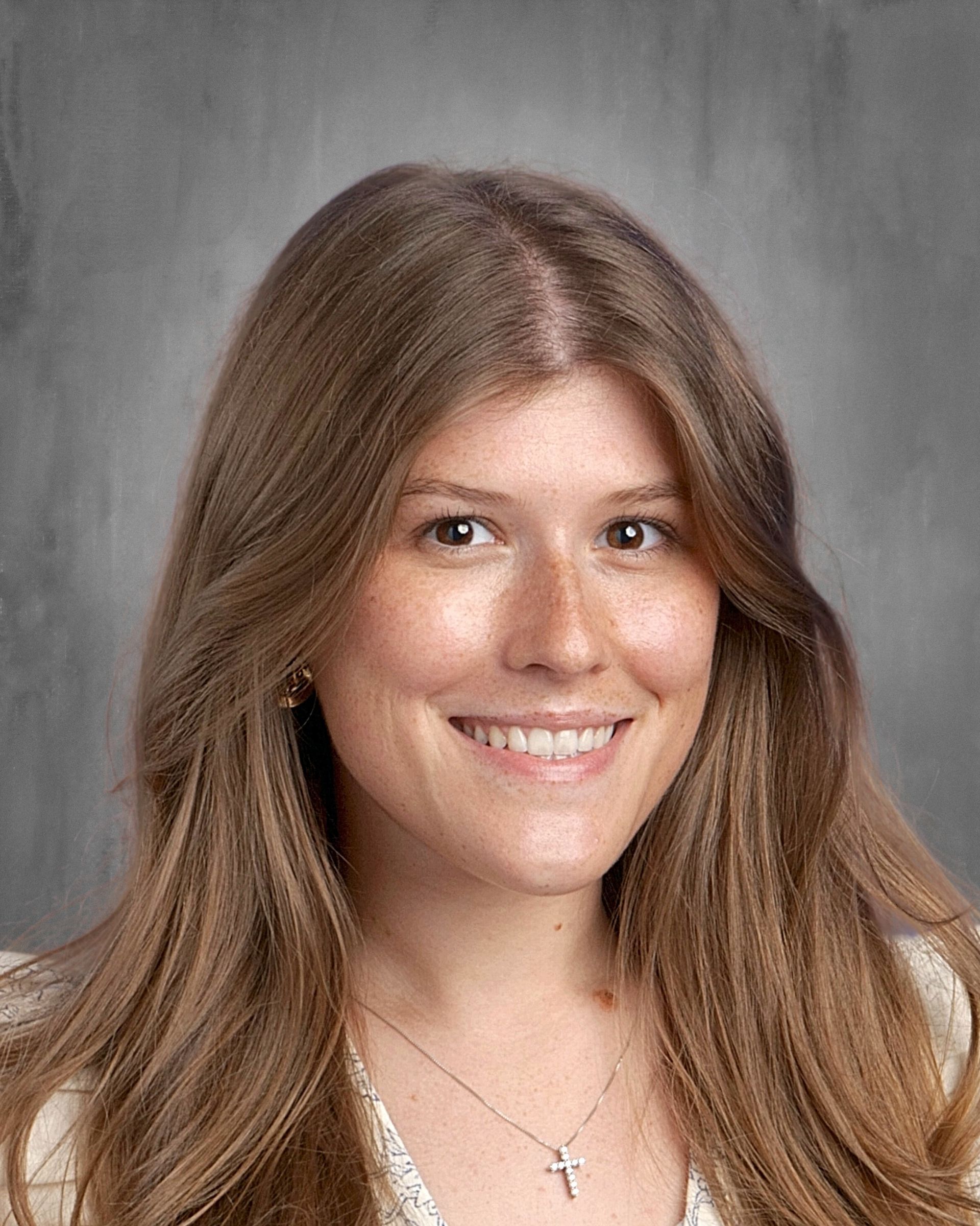 Woman with short blonde hair, glasses, smiling. Wearing a blue floral top, against a gray backdrop.