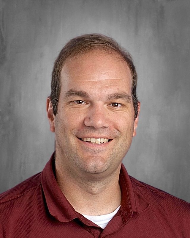 Man in a white shirt and patterned tie smiling, posed against a blurred gray backdrop.