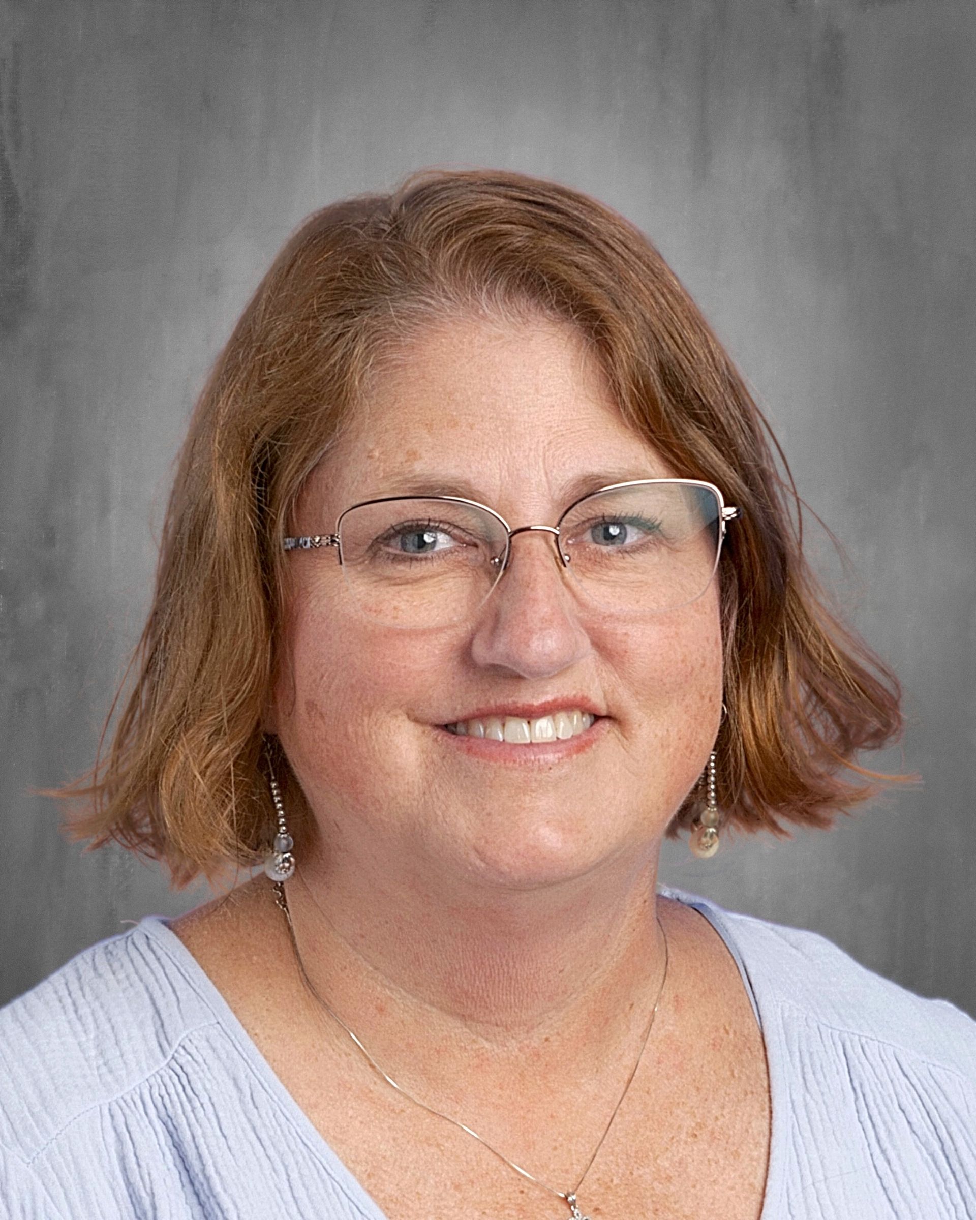 Woman with blonde hair, glasses, and a smile, wearing a light blue top. Gray background.