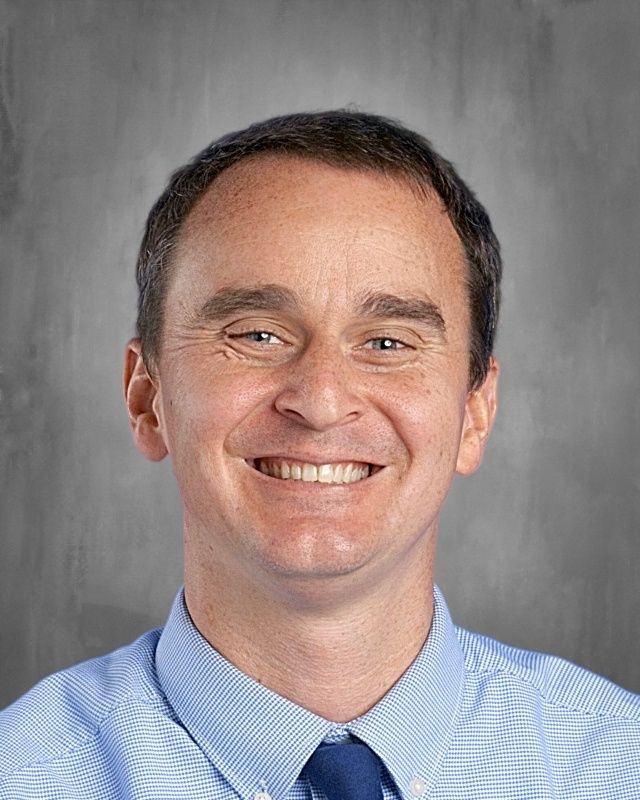 Man with short brown hair smiling, wearing a light blue collared shirt and navy tie.