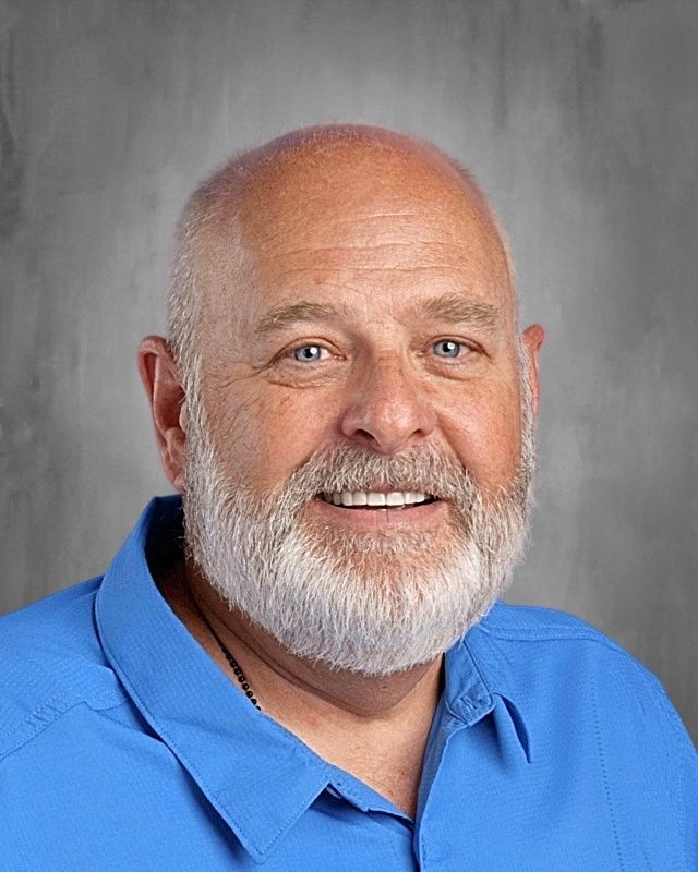 Smiling, bald man with a white beard wearing an orange polo shirt. Against a gray background.