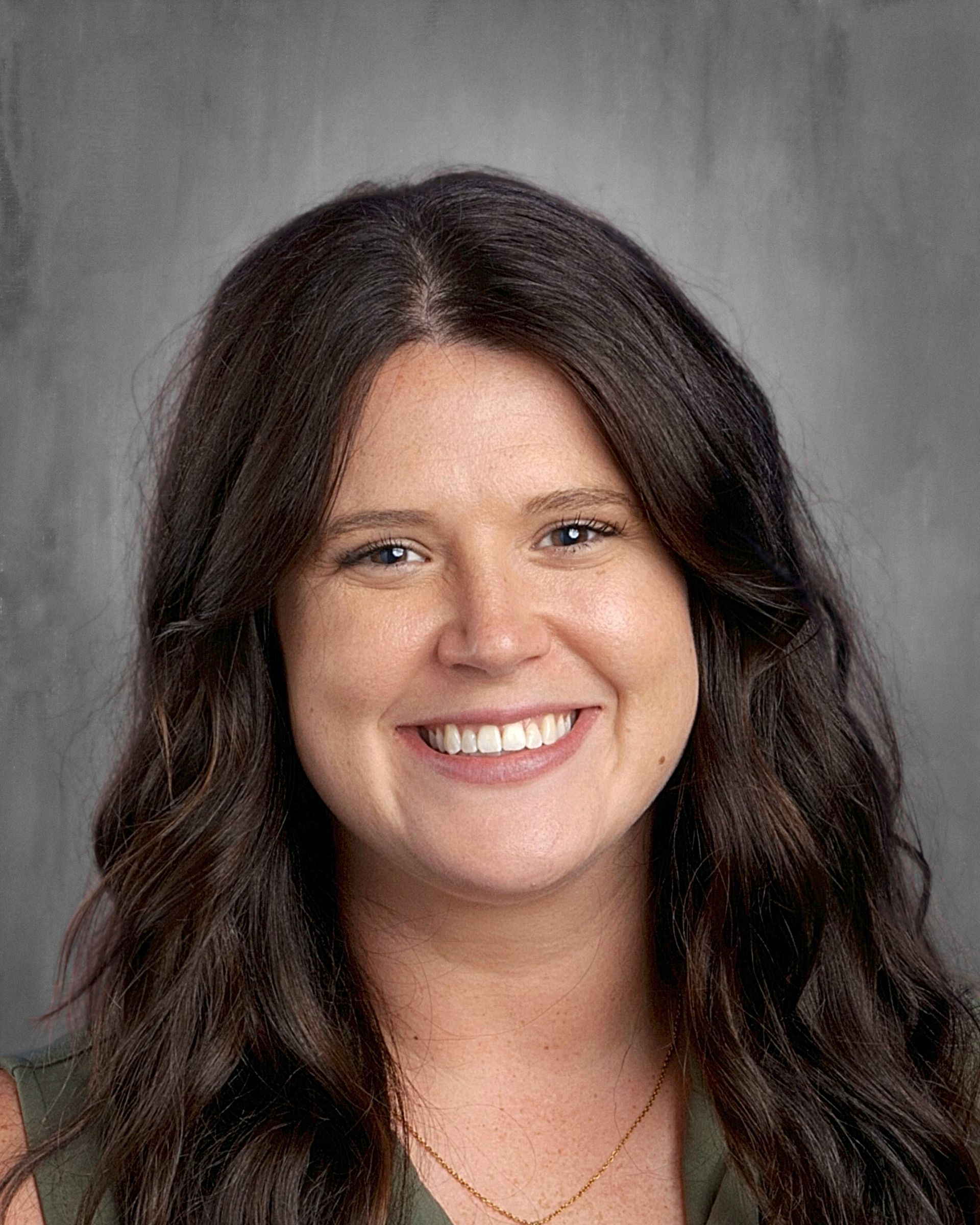 Woman with long, dark hair smiles brightly, wearing a dark top, against a gray background.