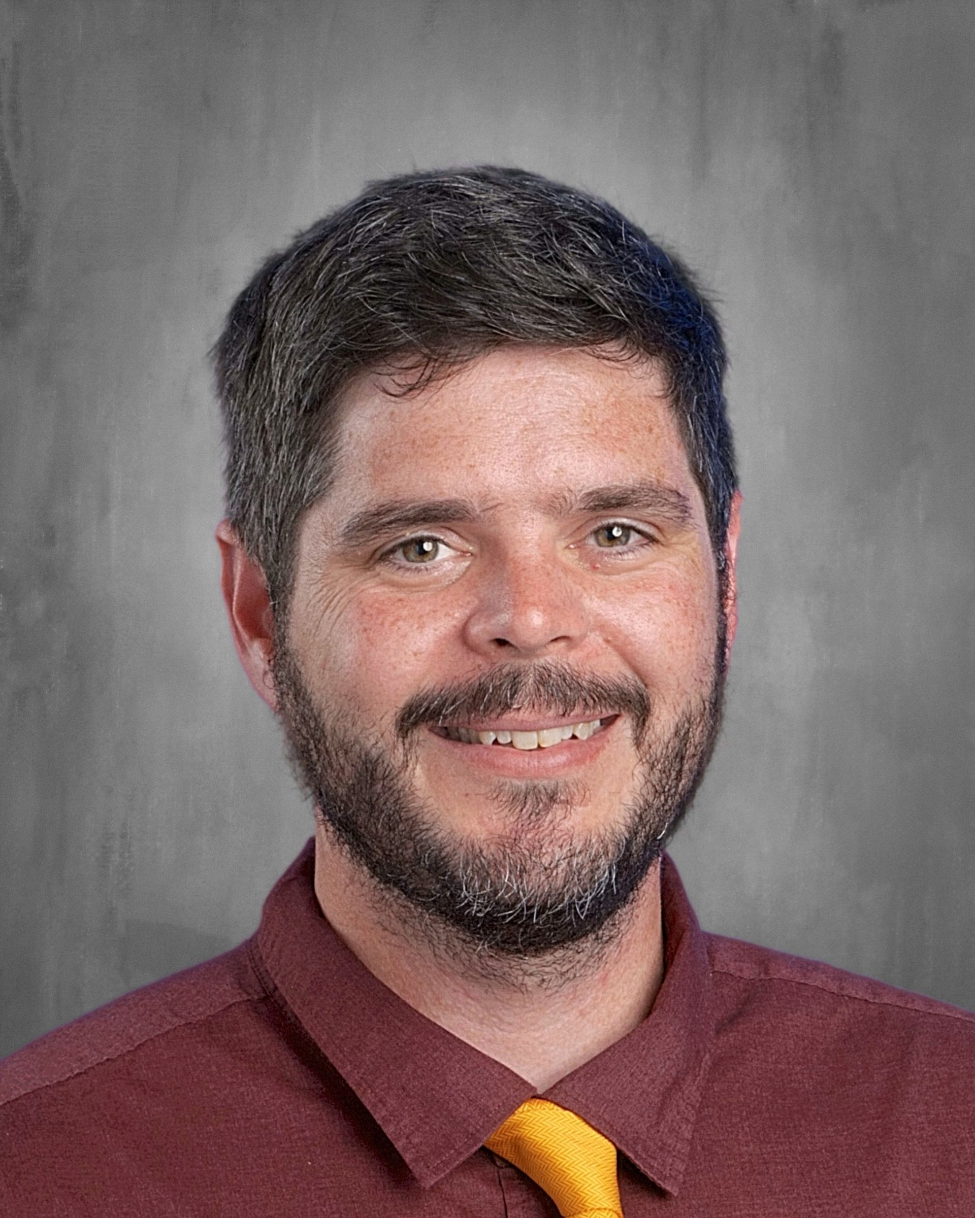 Man with dark hair and beard smiles, wearing a grey button-down shirt and tie, against a grey backdrop.