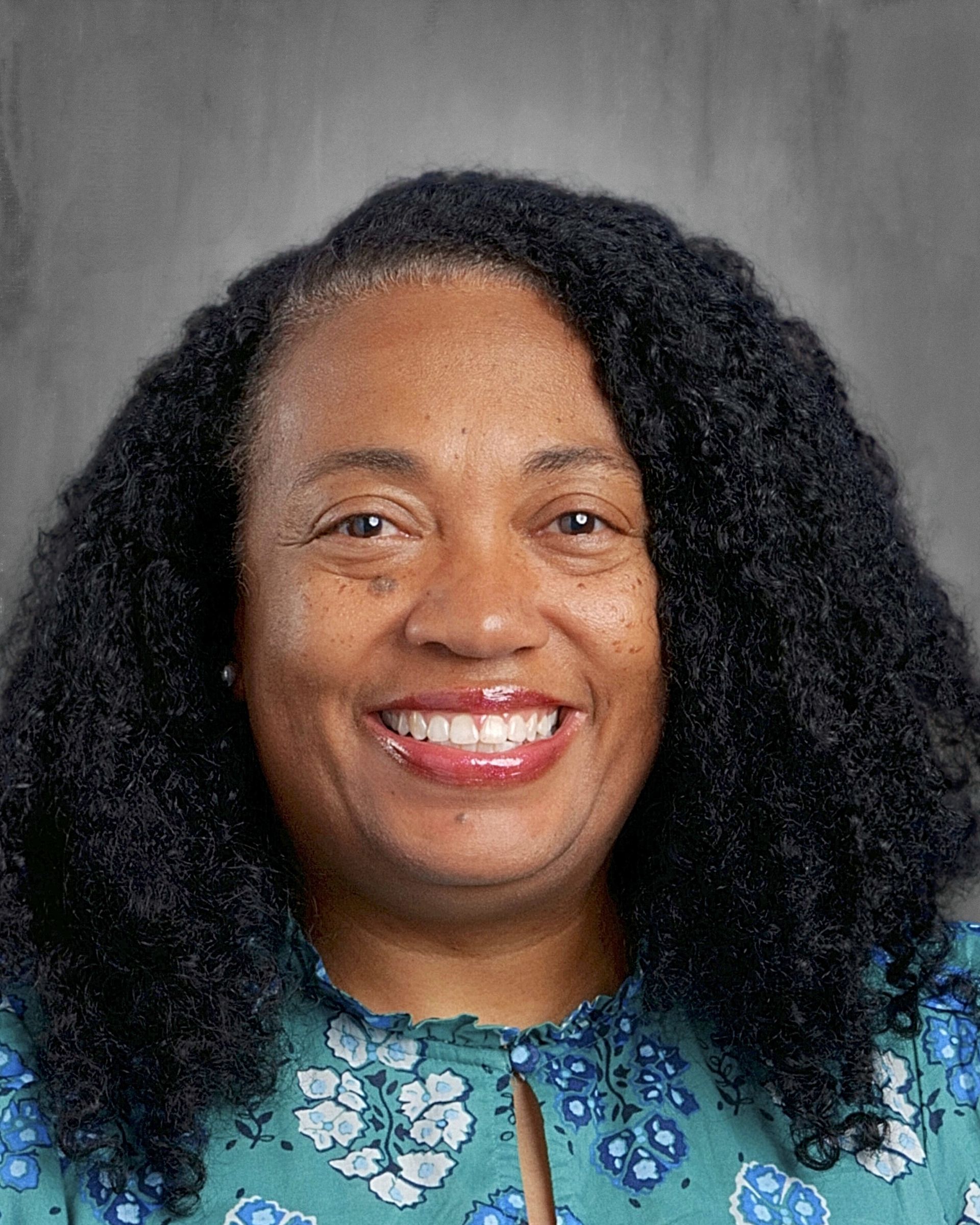 Smiling woman with curly black hair in a floral top, against a gray background.