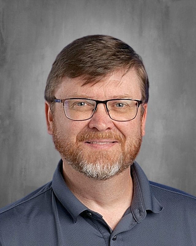 Man with glasses and beard, wearing a sweater and plaid shirt, smiles in front of a gray wall.