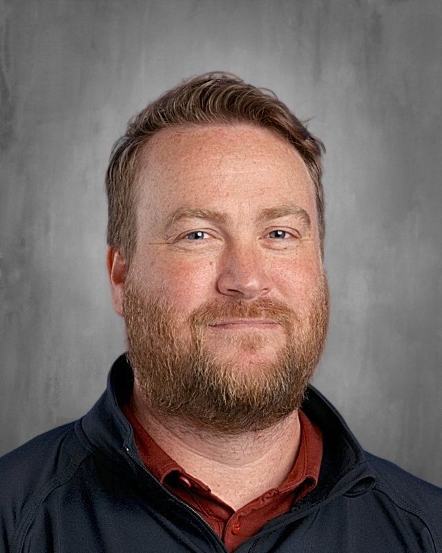Man with glasses smiles at the camera, wearing a blue polo shirt, gray background.