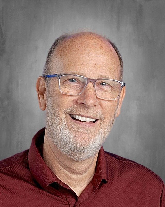 Man with glasses smiles at the camera, wearing a blue polo shirt, gray background.