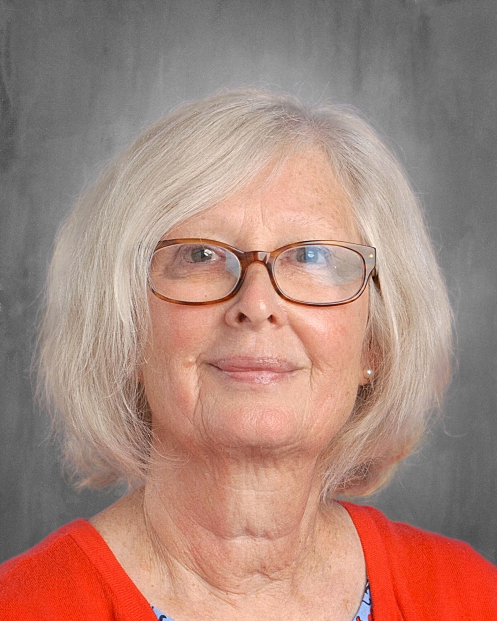 Woman with short blonde hair and glasses smiles at the camera. She wears a blue shirt, in front of a gray backdrop.