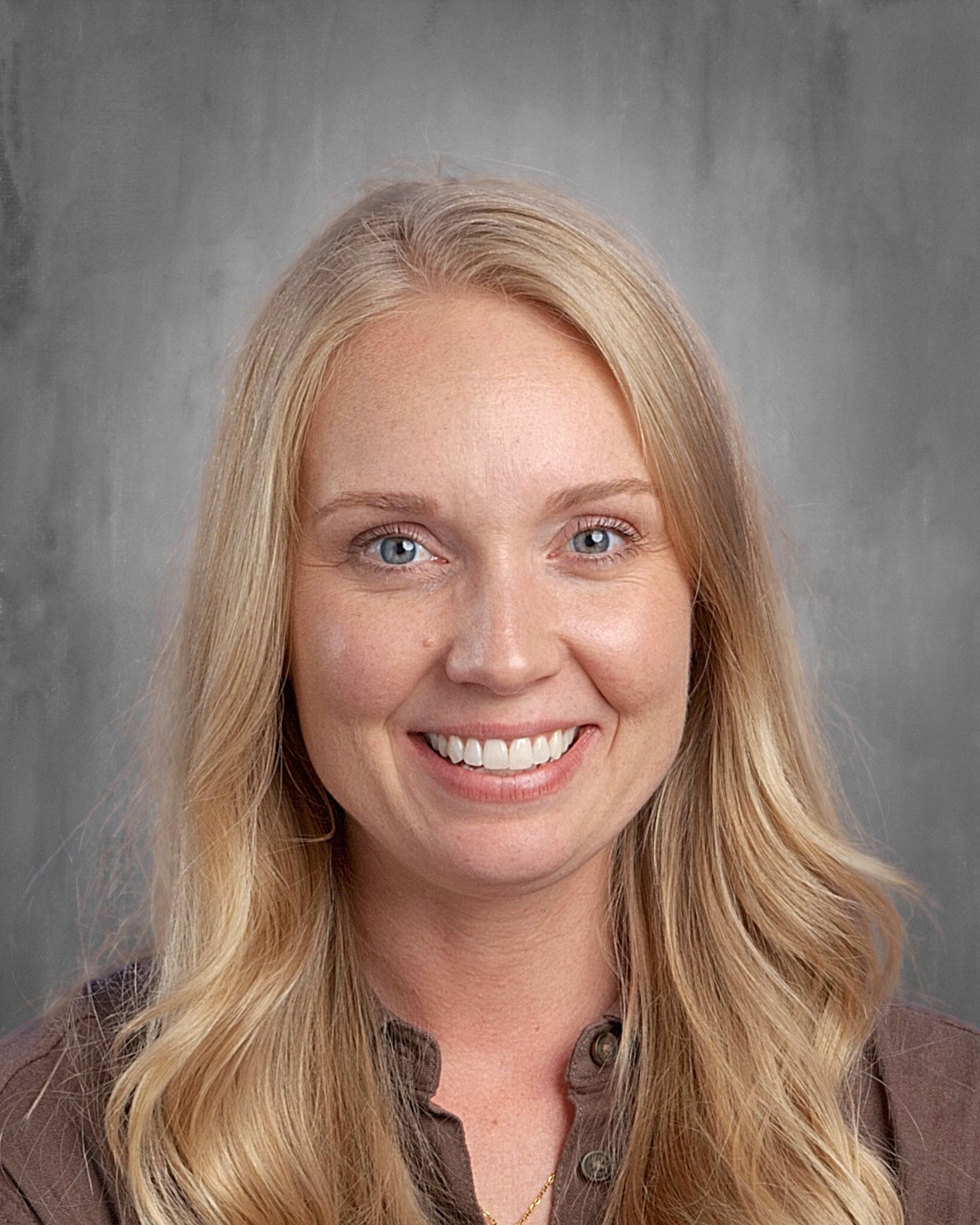 Blonde woman with blue eyes smiles, wearing a brown shirt, against a gray background.