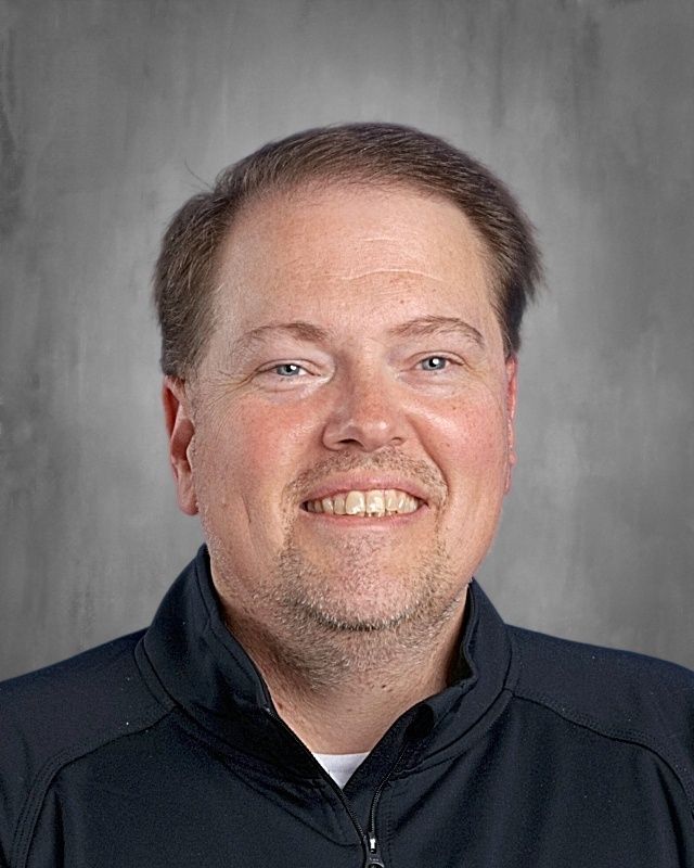 Man with fair skin wearing a collared shirt and striped tie smiles, looking at the viewer, against a grey background.