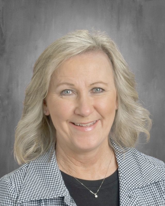 Blonde woman with blue eyes smiling, wearing a patterned jacket and necklace, against a gray background.