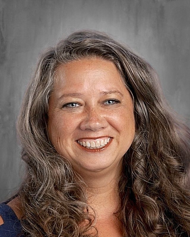 Woman with long wavy brown hair smiles at the camera; blue top, gray background.