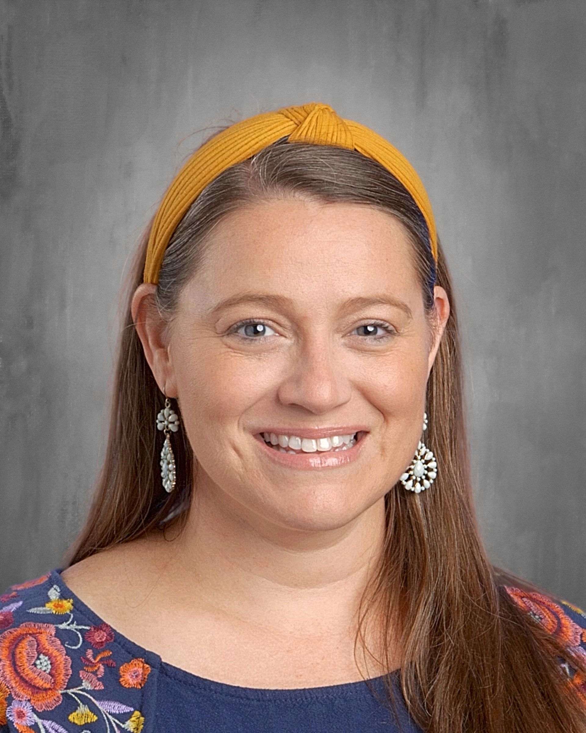 Woman with fair skin smiles, wearing flower-patterned top, white earrings, and brown hair in a ponytail.