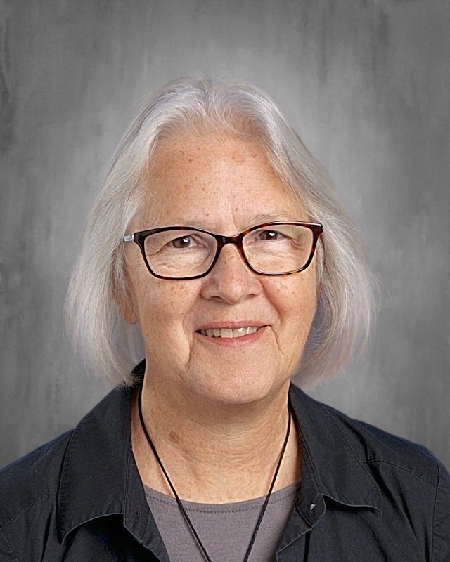Woman with gray hair and glasses smiling, wearing a blue patterned shirt.