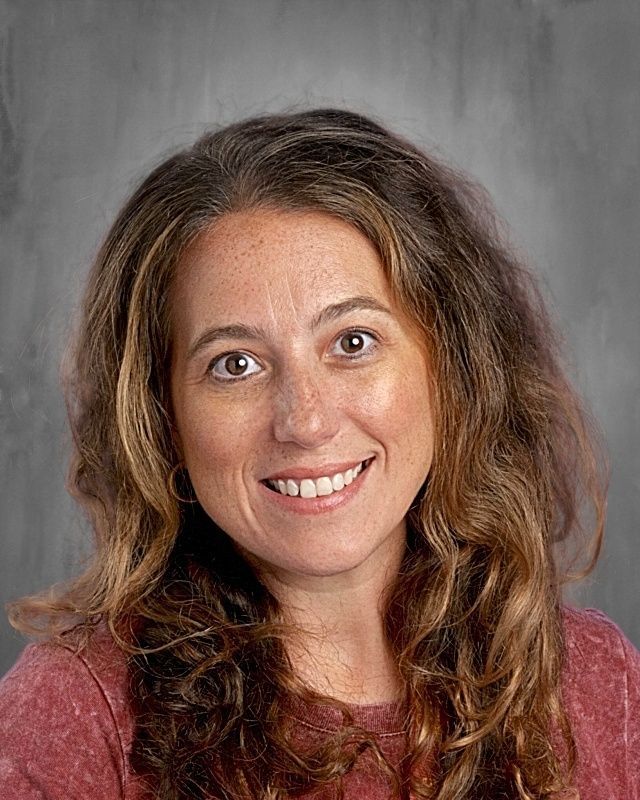 Woman with brown curly hair smiles, wearing a red shirt and a gold necklace, in front of a gray background.