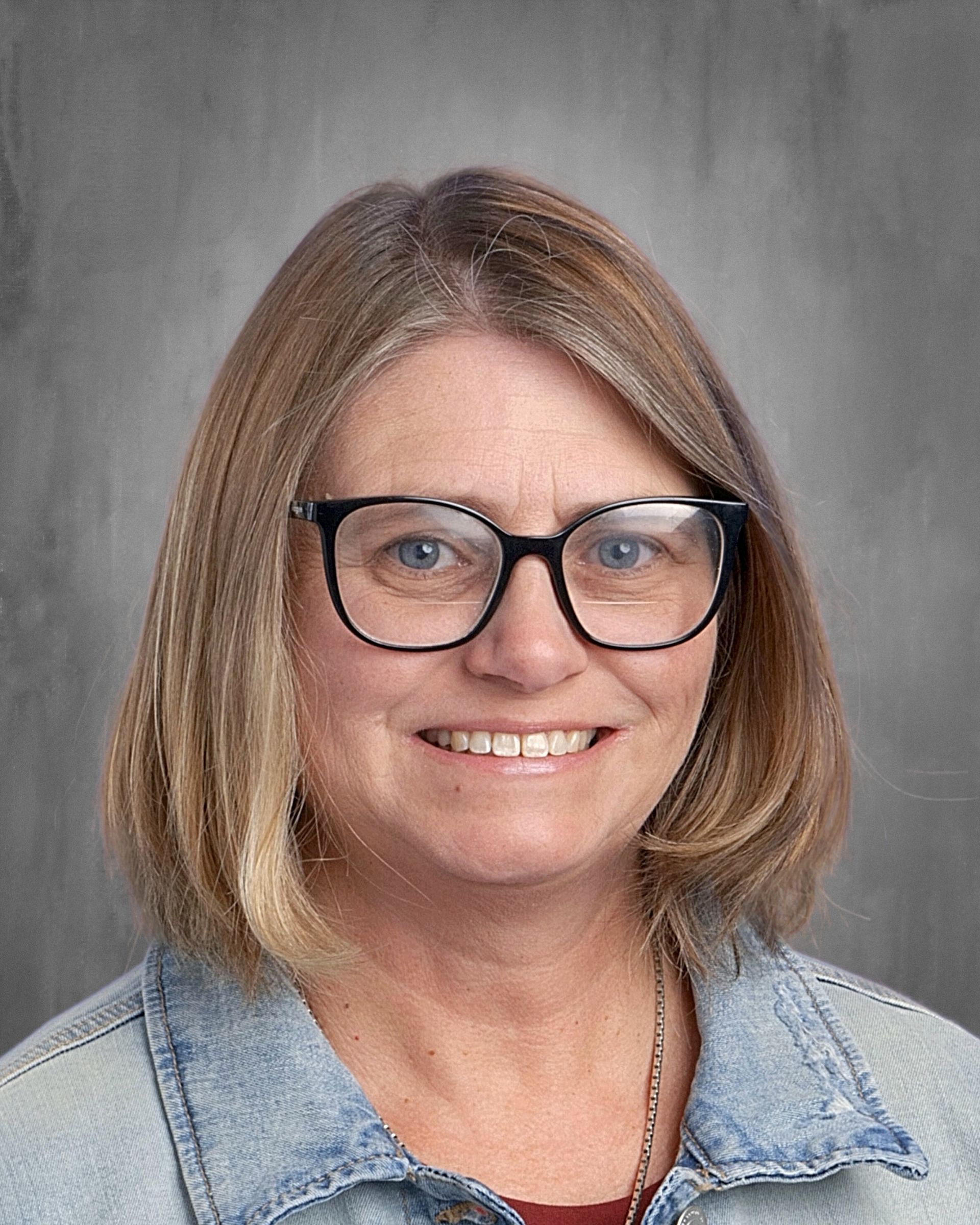 Woman with glasses smiles, wearing a dark top, in front of a gray background.