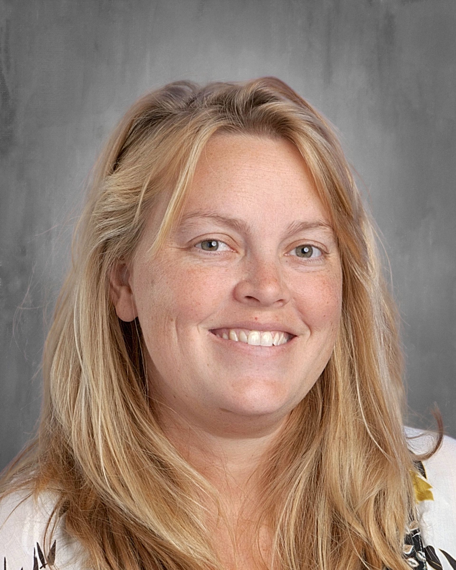 Woman with fair skin and blonde hair, smiling. She wears a floral shirt and earrings against a gray backdrop.
