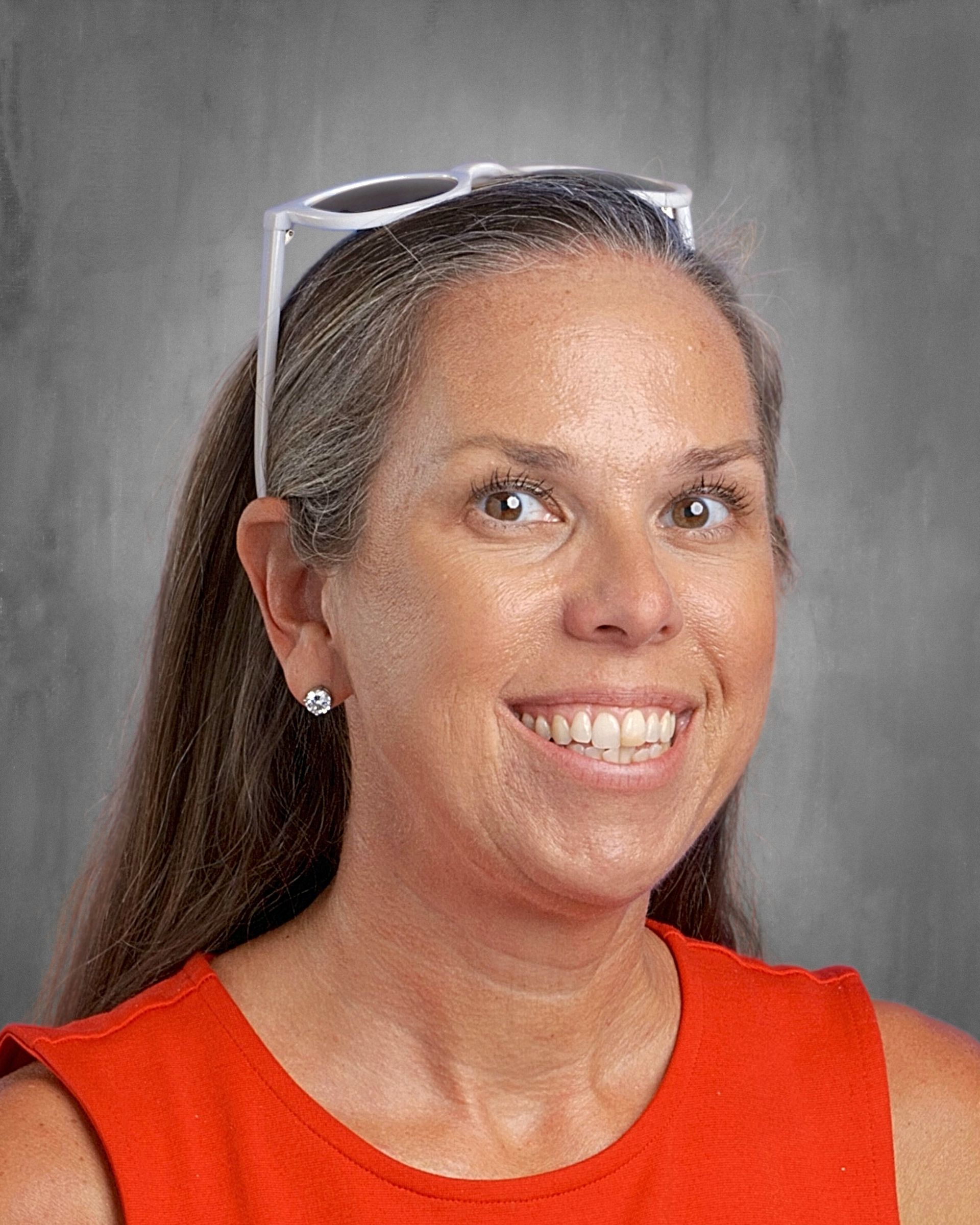 Woman with glasses on head smiles, wearing orange top, gold jewelry, against a gray background.