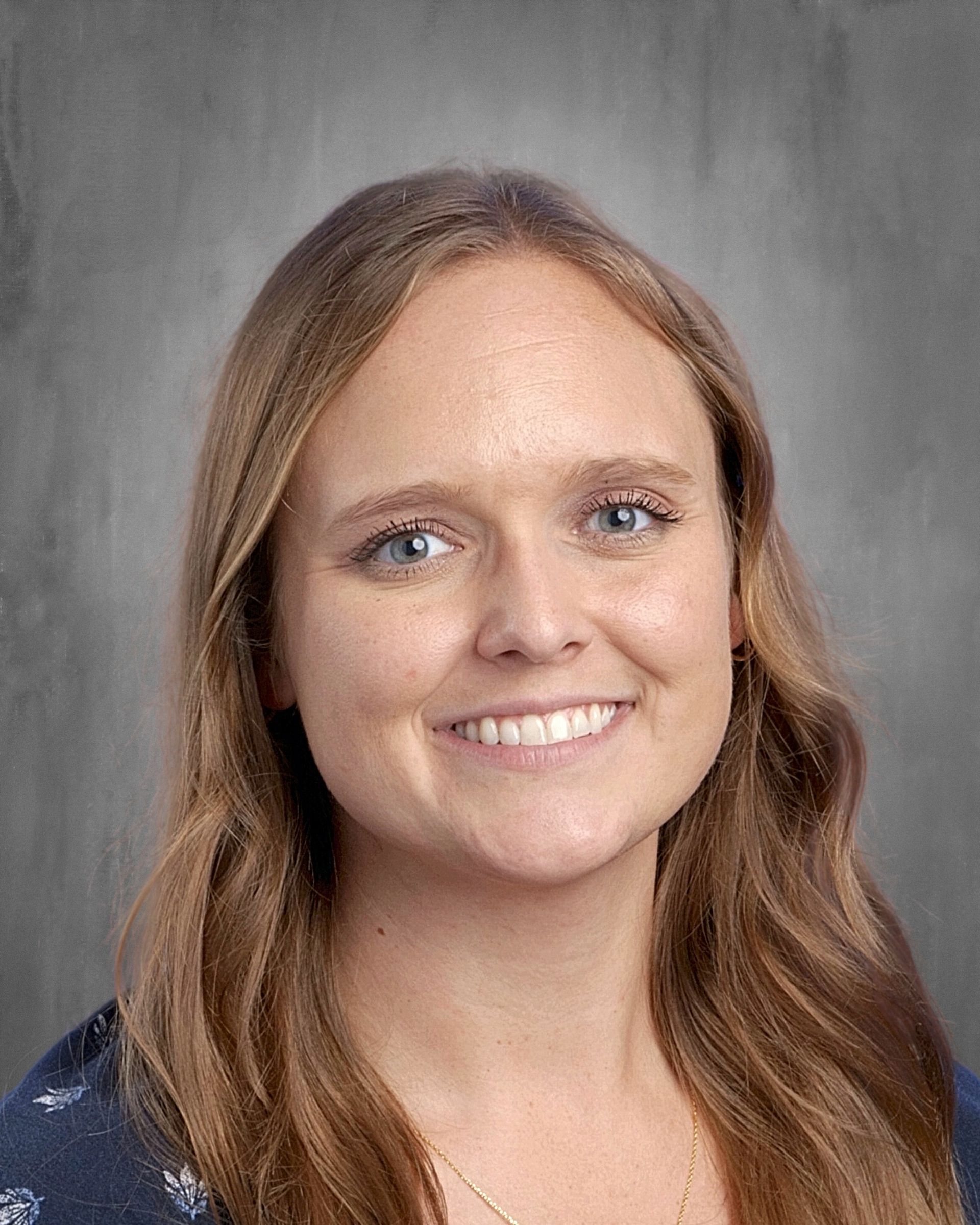 Woman with light skin, blue eyes, and shoulder-length brown wavy hair smiles at the camera.