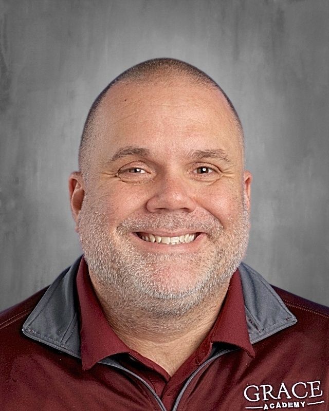 Man with shaved head smiles; wears light blue shirt and patterned tie against a gray background.