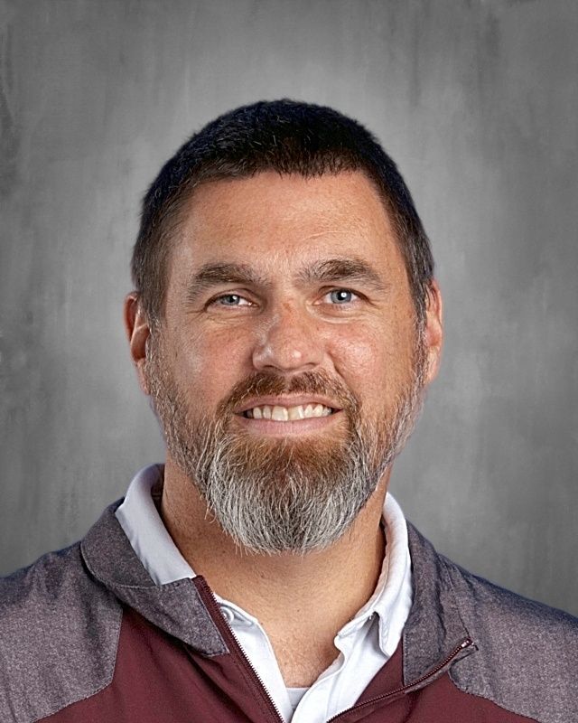 Man with beard smiles, wearing white polo shirt, against a grey backdrop.