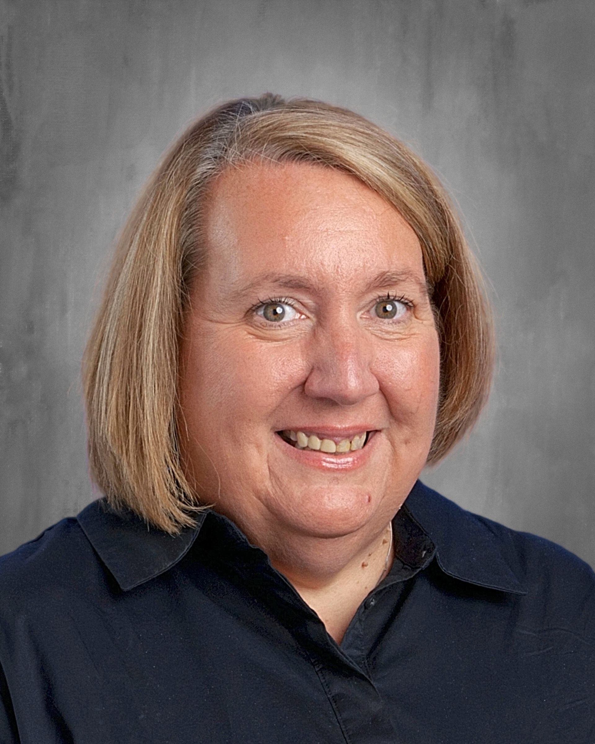 Woman with short blonde hair, smiling, wearing a blue-green patterned shirt, gray backdrop.
