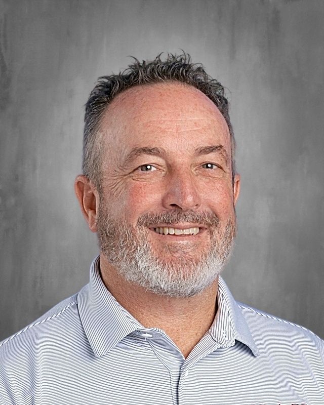 Man with a gray beard wearing a blue blazer and light blue collared shirt, smiling against a gray backdrop.